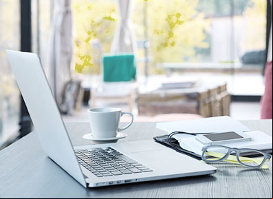 Notebook aberto posicionado em cima de uma mesa. Ao lado, uma xícara branca, um óculos de grau com a armação cinza e algumas folhas de papel