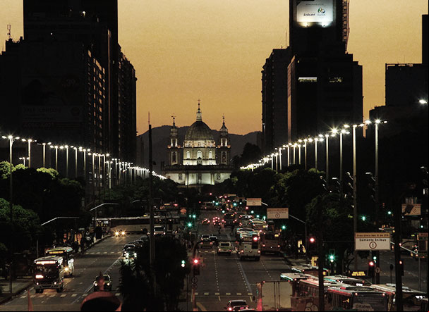 Paisagem de avenida movimentada, ao final da tarde. Nas laterais, diversos postes iluminando a rua. Ao redor, edificícios cercam a imagem.