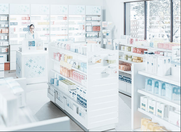 Visão interna de estabelecimento farmacéutico, com gondolas brancas e prateleiras com medicamentos.  