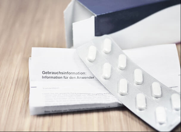 Cartela de medicamentos na cor branca, posicionados acima de uma bula. Ao fundo, caixa dos comprimidos na cor azul marinho