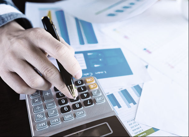 Pessoa utilizando calculadora cinza com teclas em preto e cinza. Ao lado, estão distribuidas várias folhas de papel sulfite branco com gráficos impressos
