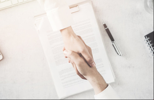 Duas pessoas brancas dando as mãos como forma de "acordo". Ao fundo da imagem, contrato assinado pelas partes em cima de uma mesa