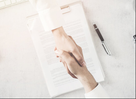 Duas pessoas brancas dando as mãos como forma de "acordo". Ao fundo da imagem, contrato assinado pelas partes em cima de uma mesa