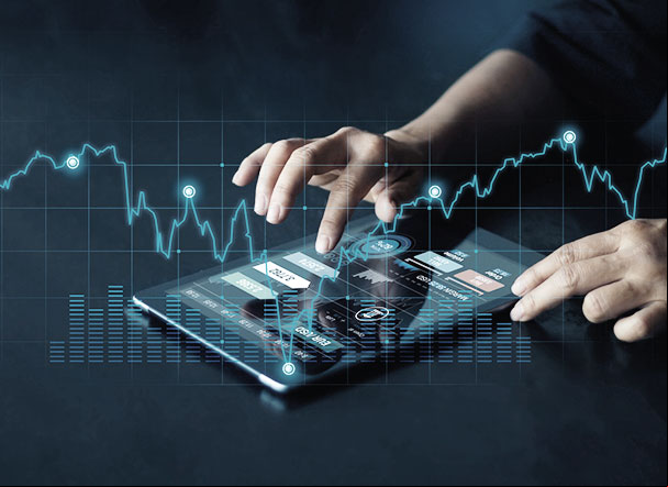 mãos de uma pessoa branca teclando em um tablet posicionado em cima de uma mesa cinza. Na tela do tablet estão informações sobre o mercado financeiro