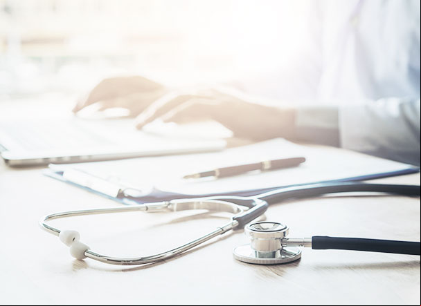 Stethoscope on a wooden table. In the background, also on the table, a clipboard with a black pen above the sheets.