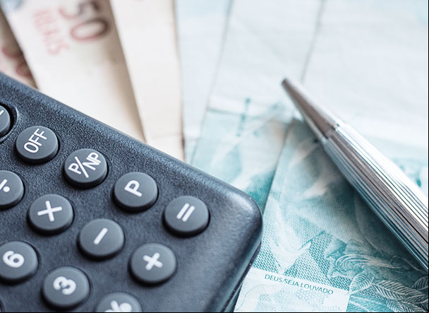 calculadora preta posicionada acima de notas de 50 e 100 reais. Do lado direito da calculadora, uma caneta prata.
