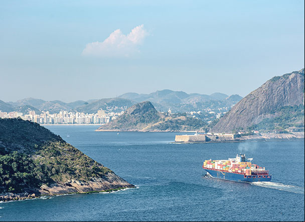 Vista superior do oceano, com navio cargueiro atravessando. Mais ao fundo, está a cidade do Rio de Janeiro